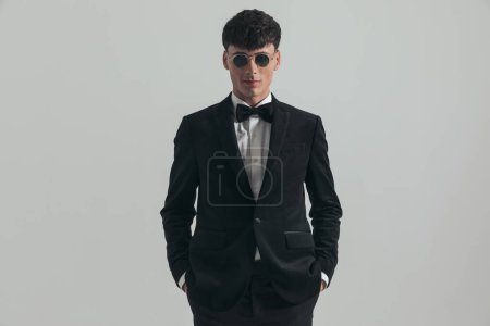 Photo for Portrait of young businessman putting his hands in pockets, standing, wearing a black tuxedo and sunglasses, in a fashion pose - Royalty Free Image