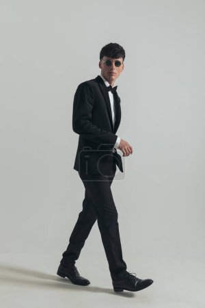 Photo for Full body picture of attractive businessman with badass walk, standing, wearing a black tuxedo and sunglasses, in a fashion pose - Royalty Free Image