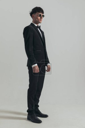 Foto de Full body picture of young businessman looking away and posing, standing, wearing a black tuxedo and sunglasses, in a fashion pose - Imagen libre de derechos