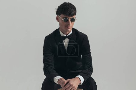 Photo for Portrait of attractive businessman rubbing his hands and looking away, sitting on a wooden chair, wearing a black tuxedo and sunglasses, in a fashion pose - Royalty Free Image