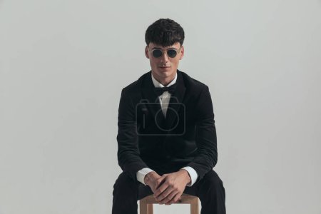 Photo for Portrait of handsome businessman rubbing his hands and smiling, sitting on a wooden chair, wearing a black tuxedo and sunglasses, in a fashion pose - Royalty Free Image