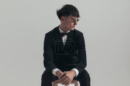 Photo for Portrait of young businessman rubbing his hands and looking to her side, sitting on a wooden chair, wearing a black tuxedo and sunglasses, in a fashion pose - Royalty Free Image
