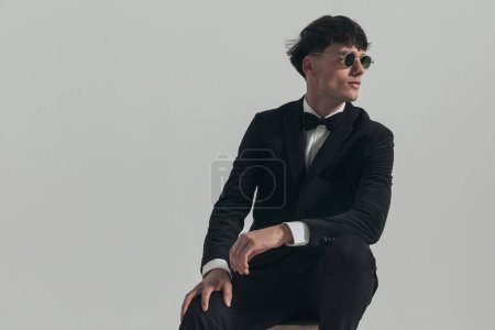 Photo for Portrait of attractive businessman resting his arms and looking to side, sitting on a wooden chair, wearing a black tuxedo and sunglasses, in a fashion pose - Royalty Free Image