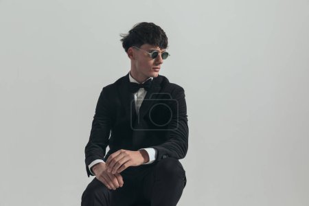 Téléchargez les photos : Portrait de jeune homme d'affaires regardant vers le bas avec une attitude dure, assis sur une chaise en bois, portant un smoking noir et des lunettes de soleil, dans une pose de mode - en image libre de droit