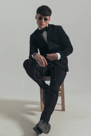 Foto de Imagen de cuerpo completo del atractivo hombre de negocios cerrando su chaqueta lentamente, sentado en una silla de madera, con un esmoquin negro y gafas de sol, en una pose de moda - Imagen libre de derechos