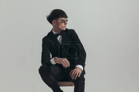 Photo for Portrait of handsome businessman posing with sexy stance, sitting on a wooden chair, wearing a black tuxedo and sunglasses, in a fashion pose - Royalty Free Image
