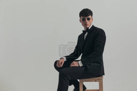 Foto de Portrait of attractive businessman posing with cool, manly vibe, sitting on a wooden chair, wearing a black tuxedo and sunglasses, in a fashion pose - Imagen libre de derechos