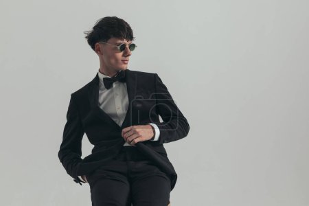Photo for Portrait of attractive businessman posing with a cool, sexy energy, sitting on a wooden chair, wearing a black tuxedo and sunglasses, in a fashion pose - Royalty Free Image