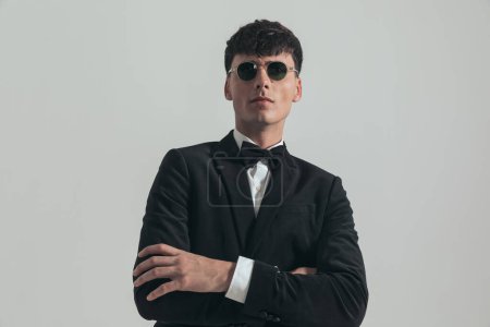 Photo for Portrait of handsome businessman crossing his arms and looking away, standing, wearing a black tuxedo and sunglasses, in a fashion pose - Royalty Free Image