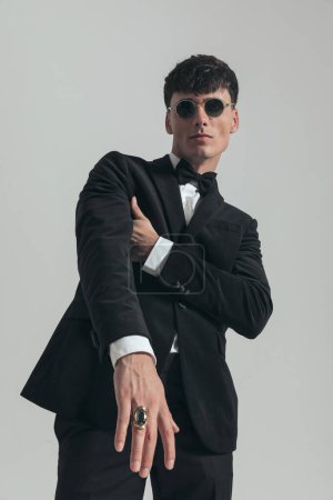 Photo for Portrait of young businessman folding one arm and showing his ring, standing, wearing a black tuxedo and sunglasses, in a fashion pose - Royalty Free Image