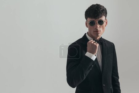Téléchargez les photos : Portrait of attractive businessman arranging his bow tie, standing, wearing a black tuxedo and sunglasses, in a fashion pose - en image libre de droit