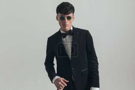 Téléchargez les photos : Portrait d'un bel homme d'affaires posant avec une position sexy, debout, portant un smoking noir et des lunettes de soleil, dans une pose de mode - en image libre de droit