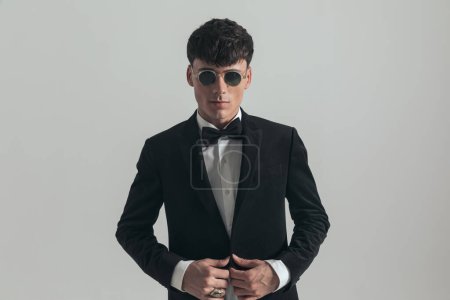 Photo for Portrait of young businessman unbuttoning his tux, standing, wearing a black tuxedo and sunglasses, in a fashion pose - Royalty Free Image