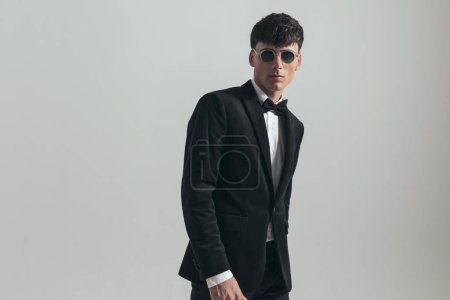 Foto de Portrait of attractive businessman posing with hands loosely around body, standing, wearing a black tuxedo and sunglasses, in a fashion pose - Imagen libre de derechos