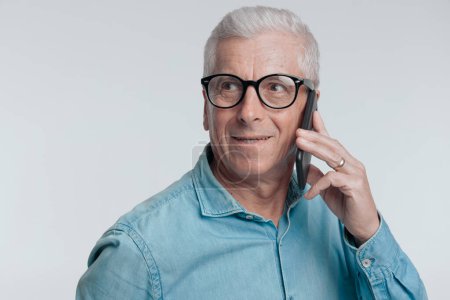 Photo for Handsome man in his 60s looking away and talking on the phone in front of grey background - Royalty Free Image