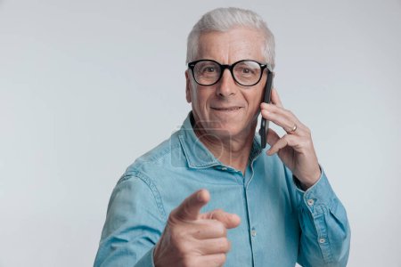 Téléchargez les photos : Portrait d'homme dans la soixantaine ayant une conversation téléphonique, souriant et pointant du doigt sur fond gris en studio - en image libre de droit