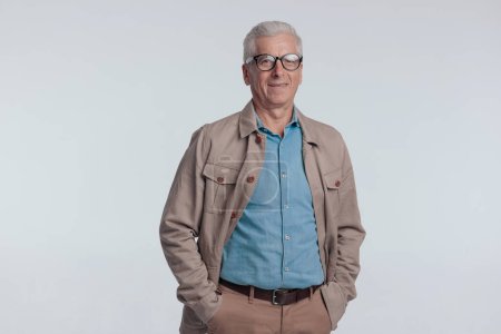 Foto de Old man in casual outfit with beige jacket smiling and holding hands in pockets on grey background - Imagen libre de derechos