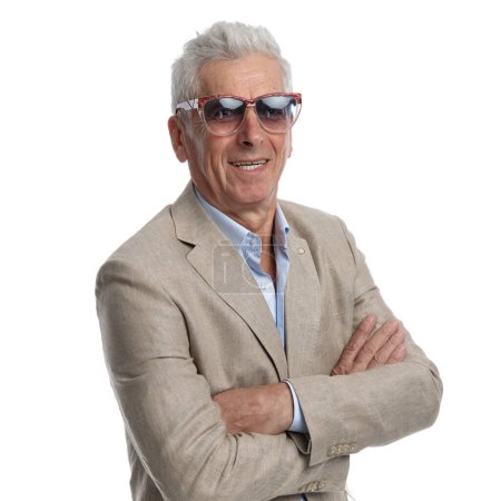 Photo for Fashion man in his 60s in elegant suit with open collar shirt wearing sunglasses, smiling and crossing arms on white background - Royalty Free Image