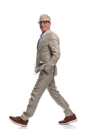 Foto de Feliz hombre de 60 años cogido de la mano en los bolsillos, sonriendo y caminando frente al fondo blanco en el estudio - Imagen libre de derechos