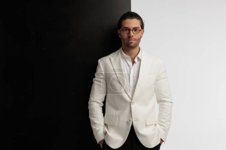 Téléchargez les photos : Portrait d'homme d'affaires attrayant mettant les mains dans les poches, debout, portant des lunettes sur fond noir et gris - en image libre de droit