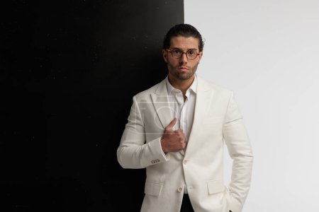 Téléchargez les photos : Portrait de beau modèle masculin arrangeant son collier avec une main dans la poche, debout, portant des lunettes sur fond noir et gris - en image libre de droit