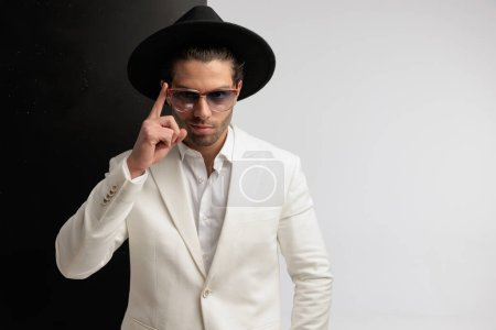 Photo for Portrait of young man fixing his eyeglasses and smiling, standing, wearing a black hat against black and gray background - Royalty Free Image
