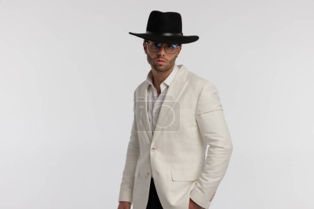 Foto de Retrato de atractivo hombre casual inteligente con buen estilo es posar, de pie, con gafas y sombrero negro sobre fondo gris - Imagen libre de derechos