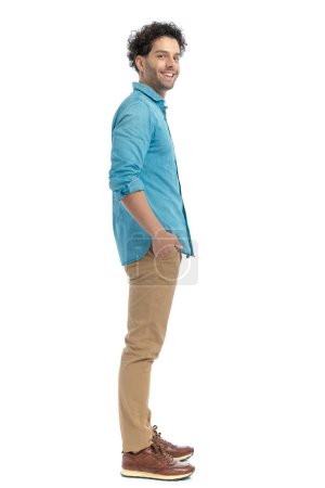Foto de Attractive casual guy with curly hair holding hands in pockets and smiling while standing in line in front of white background in studio - Imagen libre de derechos