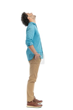 Photo for Side view of curly man with hands in pockets looking up at the sky and smiling in front of white background - Royalty Free Image