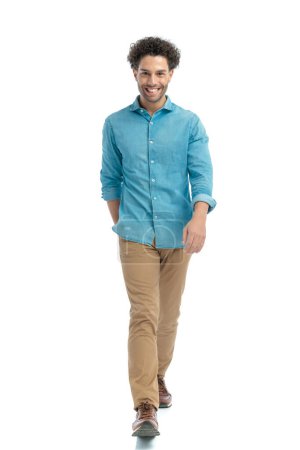 Téléchargez les photos : Happy turkish man with curly hair in denim shirt and chino pants walking and smiling in front of white background in studio - en image libre de droit