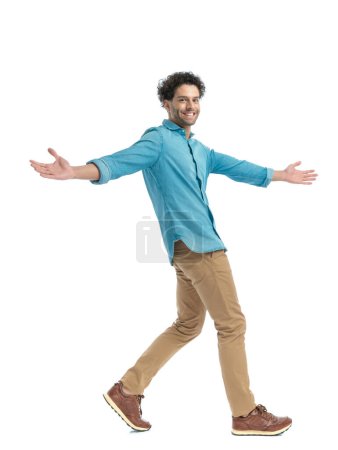 Foto de Full body picture of happy arab man in denim shir topening arms, smiling and walking in front of white background in studio - Imagen libre de derechos