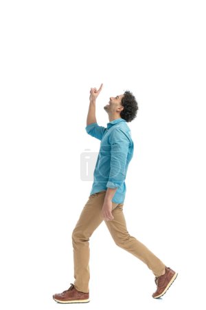 Photo for Side view of thankful young man pointing finger to the sky, walking and smiling in front of white background - Royalty Free Image