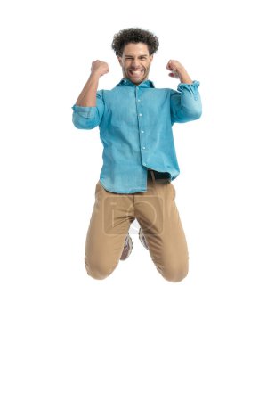 Photo for Excited turkish man with curly hair jumping in the air and cheering the victory with fists in the air in front of white background in studio - Royalty Free Image