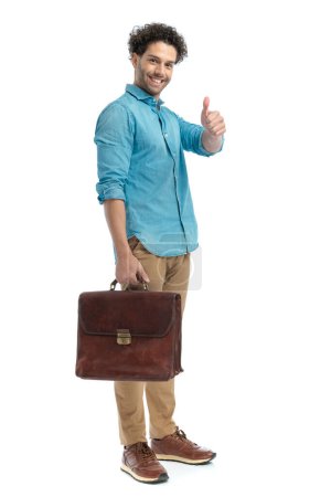 Téléchargez les photos : Fier jeune homme faisant le pouce vers le haut geste, tenant la valise et souriant devant fond blanc en studio - en image libre de droit