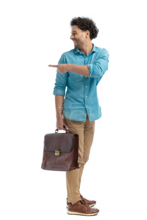 Téléchargez les photos : Side view of curly hair man looking behind, pointing finger and smiling while standing and holding bag in front of white background - en image libre de droit