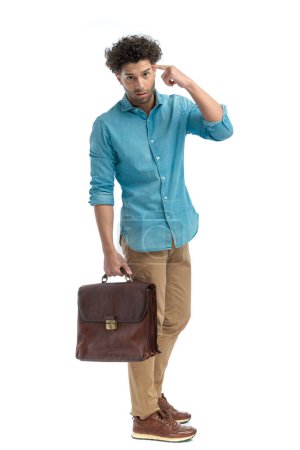 Foto de Upset young man holding suitcase and pointing finger to temple while standing on white background in studio - Imagen libre de derechos