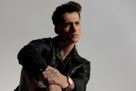 Téléchargez les photos : Pensive young man in leather jacket holding elbow on knee and looking away while thinking in front of grey background in studio - en image libre de droit