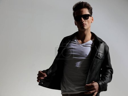 Téléchargez les photos : Sexy fashion man with sunglasses adjusting black leather jacket and posing in front of grey background in studio - en image libre de droit