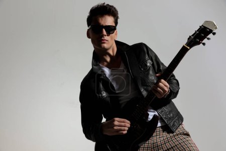 Photo for Portrait of cool guitarist with leather jacket and sunglasses playing guitar in front of grey background - Royalty Free Image