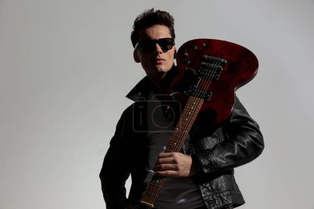 Téléchargez les photos : Sexy cool guy avec des lunettes de soleil portant la guitare sur son épaule et regardant loin devant fond gris - en image libre de droit