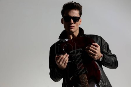 Téléchargez les photos : Sexy cool homme portant une veste en cuir noir et tenant la guitare tout en posant devant fond gris en studio - en image libre de droit