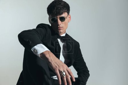 Téléchargez les photos : Élégant jeune homme d'affaires en smoking noir tenant le coude sur le genou et posant devant fond gris - en image libre de droit