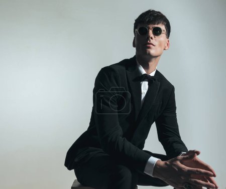 Photo for Attractive elegant groom with glasses looking up, touching palms and dreaming about his future life while sitting on grey background - Royalty Free Image