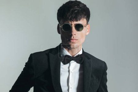 Téléchargez les photos : Portrait of sexy elegant man in black tuxedo with sunglasses posing in front of grey background - en image libre de droit