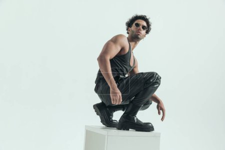 Téléchargez les photos : Handsome casual man squatting on the chair and flexing his muscles, wearing a leather costume in a fashion pose - en image libre de droit