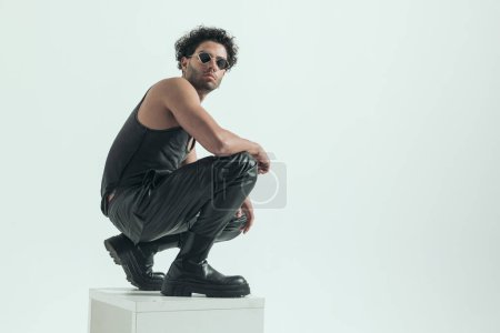 Téléchargez les photos : Attractive casual man squatting on the chair and posing, wearing a leather costume in a fashion pose - en image libre de droit