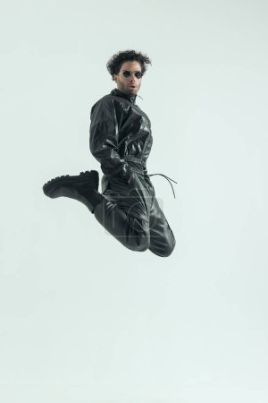 Foto de Atractivo hombre casual saltando y pateando con las manos en los bolsillos, usando un traje de cuero en una pose de moda - Imagen libre de derechos
