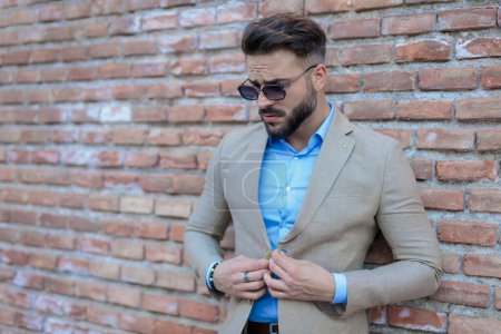 Foto de Sexy bearded man with sunglasses buttoning suit and looking down while posing outside in front of bricks wall background - Imagen libre de derechos