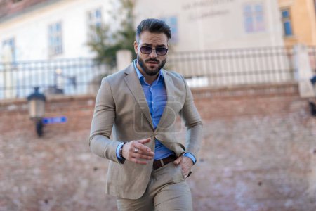 Téléchargez les photos : Sexy homme non rasé avec des lunettes de soleil marche avec la main dans la poche en plein air dans une vieille ville de Transylvanie - en image libre de droit