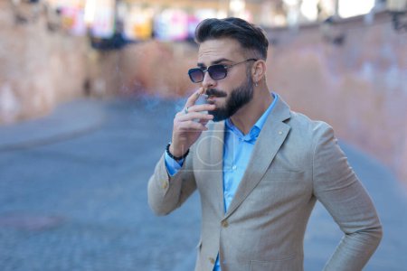 Foto de Sexy hombre de negocios en traje con gafas de sol mirando a un lado mientras fuma fuera en una ciudad vieja de Rumania - Imagen libre de derechos